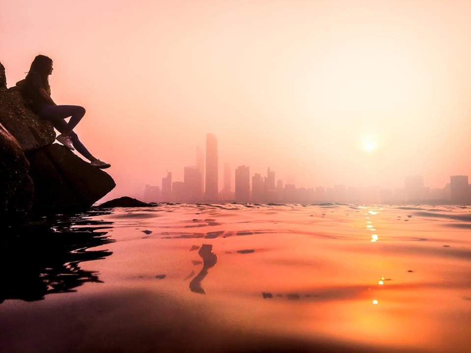 At sunset, a person sitting on a rock next to the water is in silhouette. The towers of Abu Dhabi City are in the distance.