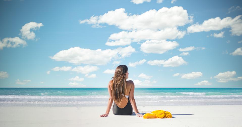 A woman with long hair wearing a black and white bathing suits sits on the white sand beach with her back to the camera. In the background is the blue-green sea and a blue sky with fluffy white clouds.
