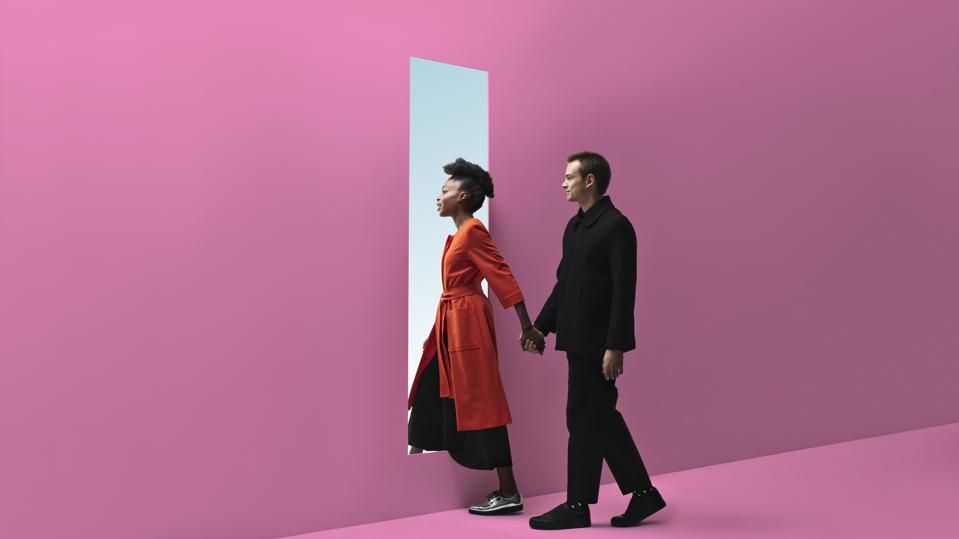 Woman & man holding hands, approaching rectangular opening in coloured wall