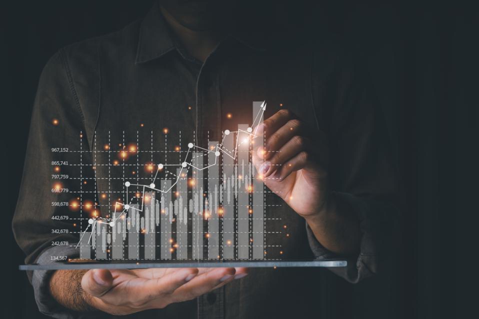 Shadowed photo of person holding digital tablet that has a digital rising business graph over it. The person is touching the tallest bar in the graph. CTOs guiding business growth concept.