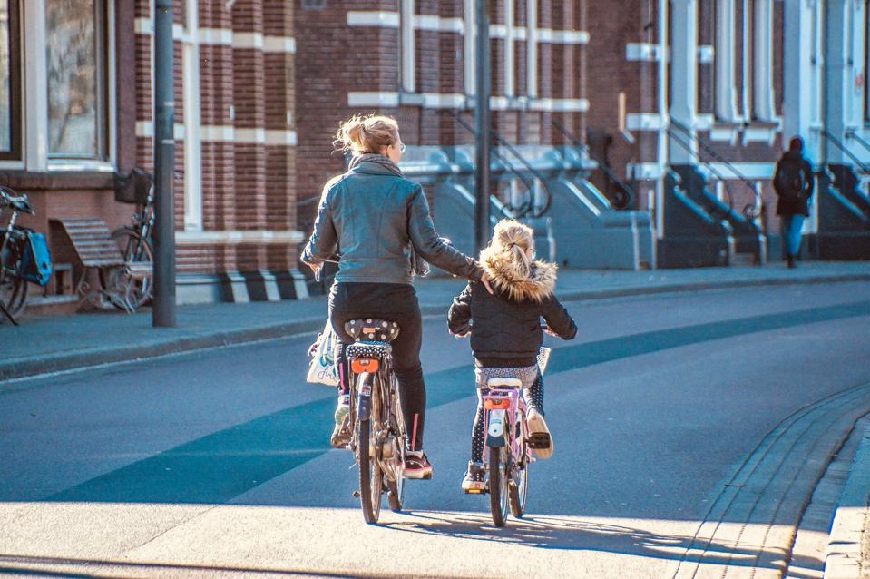 A mother and her young daughter, each on bikes, are seen cycling down a paved pathway
