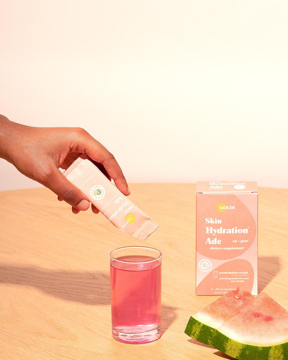 A hand pours Golde Skin Hydration Ade into a cup of water next to watermelon. 