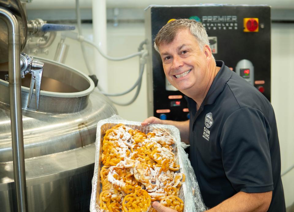 A man smiles and is holding a tray of funnel cakes before he tosses them into a beer mash.