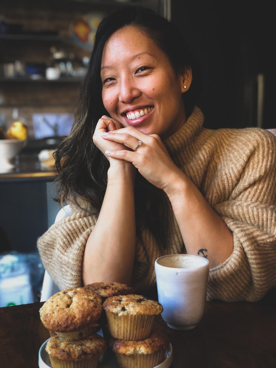Joanne Molinaro smiles with a stack of muffins in a tan sweater