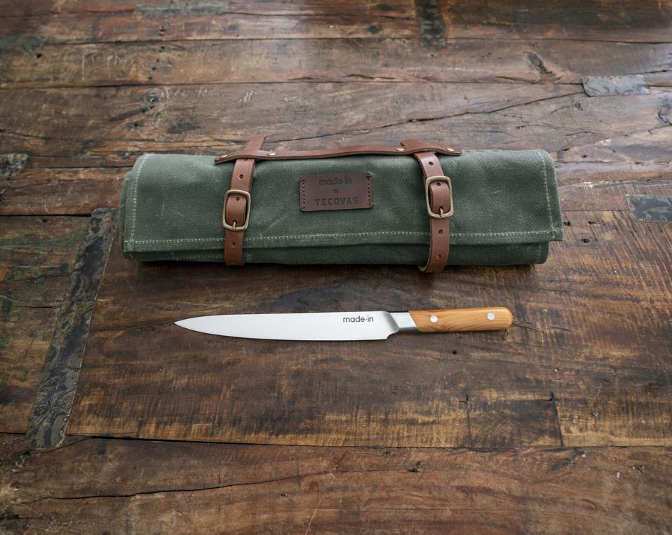 Made In and Tecovas collaborated to create an outdoor cooking adventure kit that includes a green waxed-twill knife roll with saddle-color harness leather buckles and a 9-inch carving knife with an olive wood handle that is seen in this photograph.