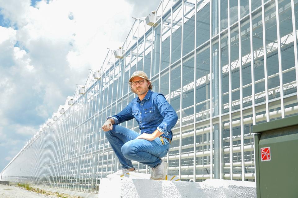 A casually dressed man crouches in front of a huge greenhouse