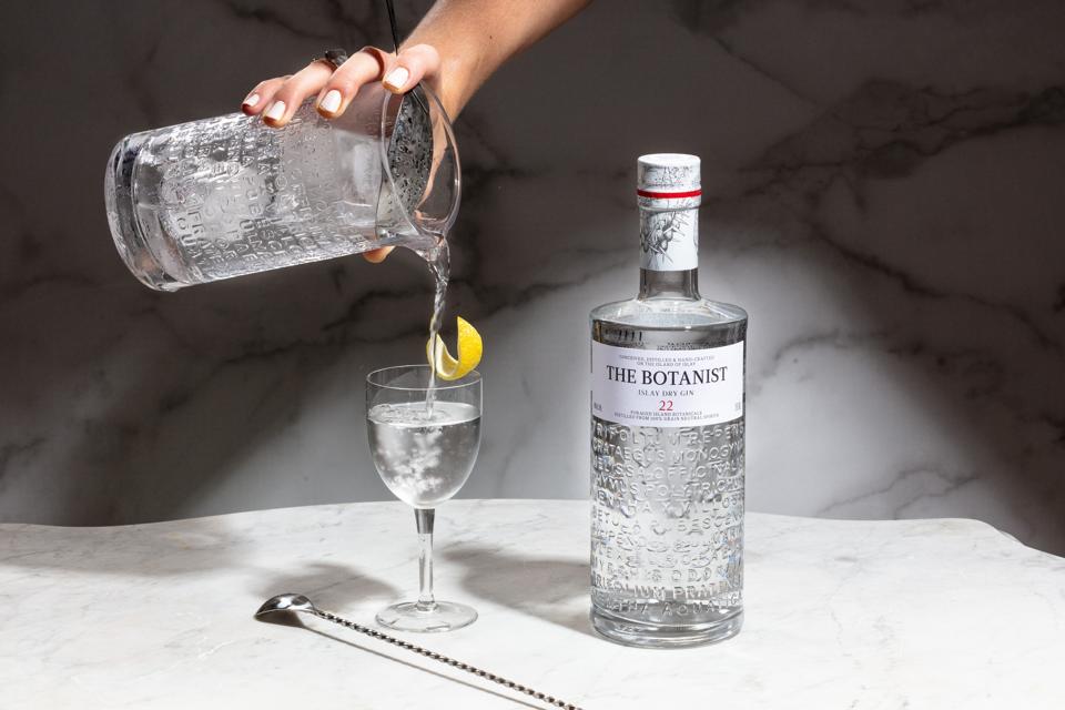 gin cocktail being poured into a glass with a bottle of gin and a stirrer next to it with a lemon peel