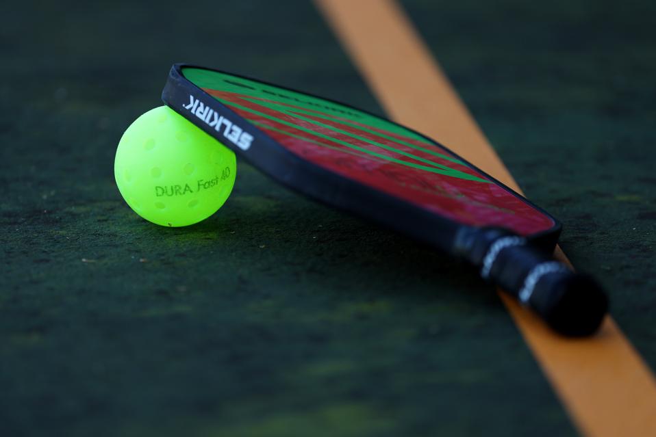 A pickleball paddle rests atop a pickleball ball on a pickkleball court.