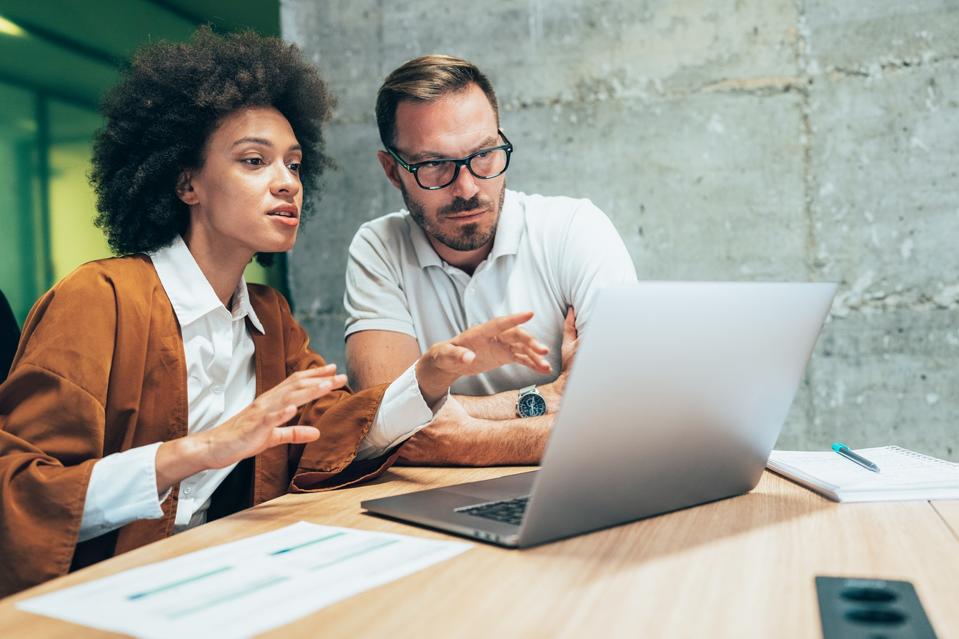 Two colleagues, male and female, looking at a laptop. The female colleague is explaining her workflow needs for new software