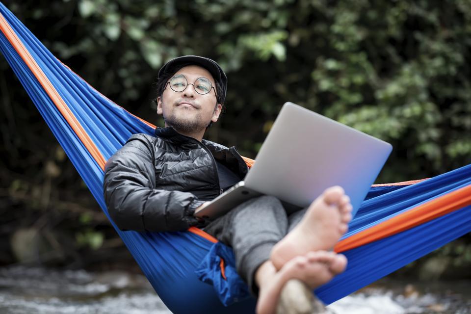 Senior Man lying in hammock using a laptop on a vacation. Camping Lover, Nature Addicted, and Remote Location.
