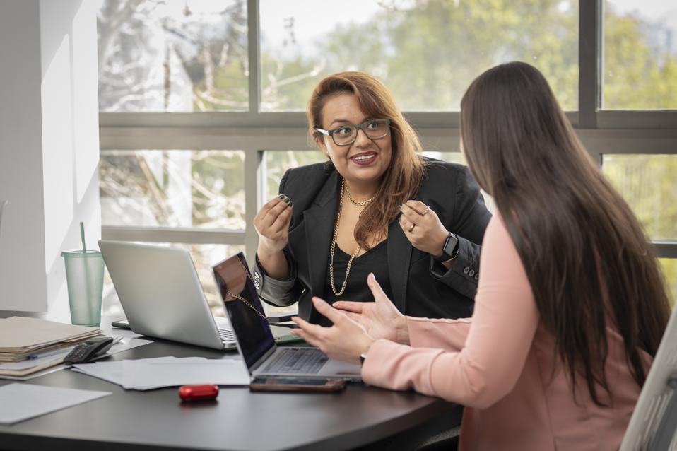 Two Latina women chat happily as they work on their laptops, sitting at a desk on an ordinary office day.