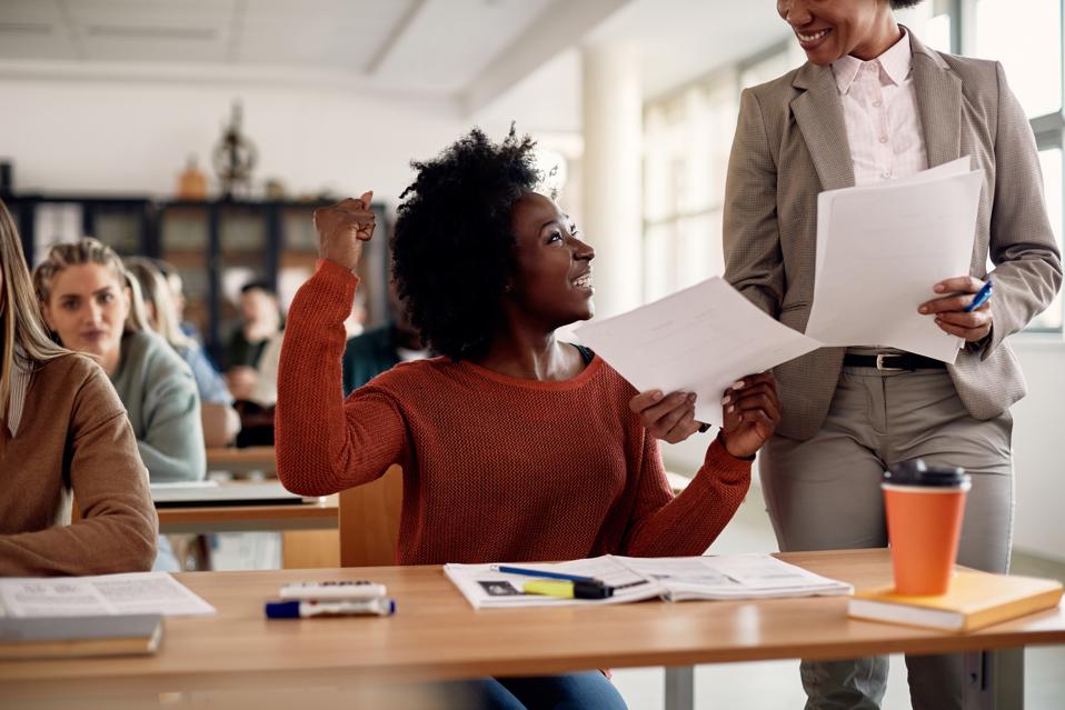 Feliz estudiante afroamericana celebrando mientras recibe los resultados de los exámenes de su profesor en el aula.