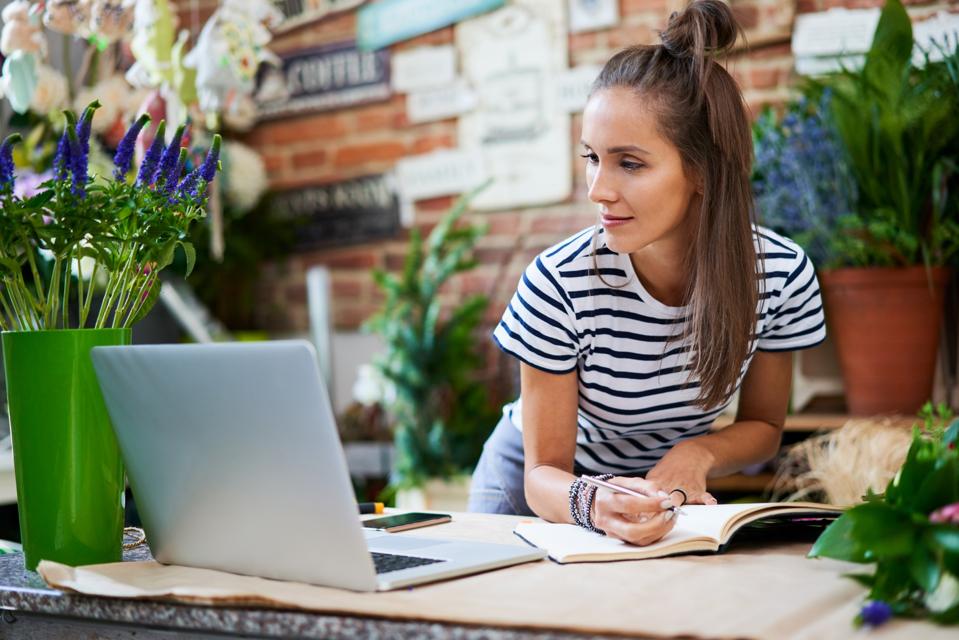 Joven florista escribiendo en un cuaderno y mirando la computadora portátil mientras se apoya en el mostrador