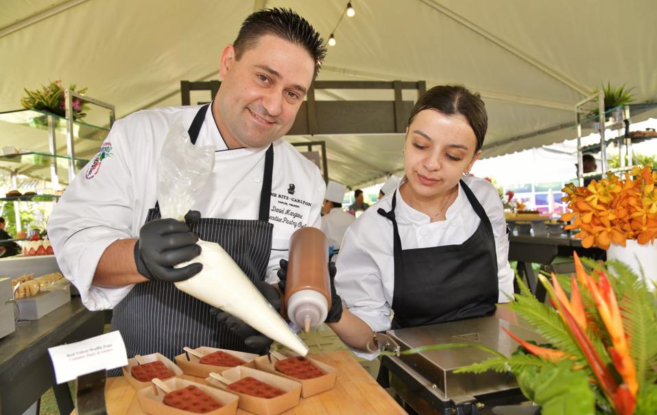 A chef with pastry cream and caramel sauce tops off desserts as another chef looks on.
