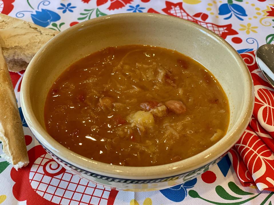 A close-up photo of a bowl of the Slovenian cabbage stew is shown in a ceramic bowl surrounded by Slovenian linens and a crusty baguette.