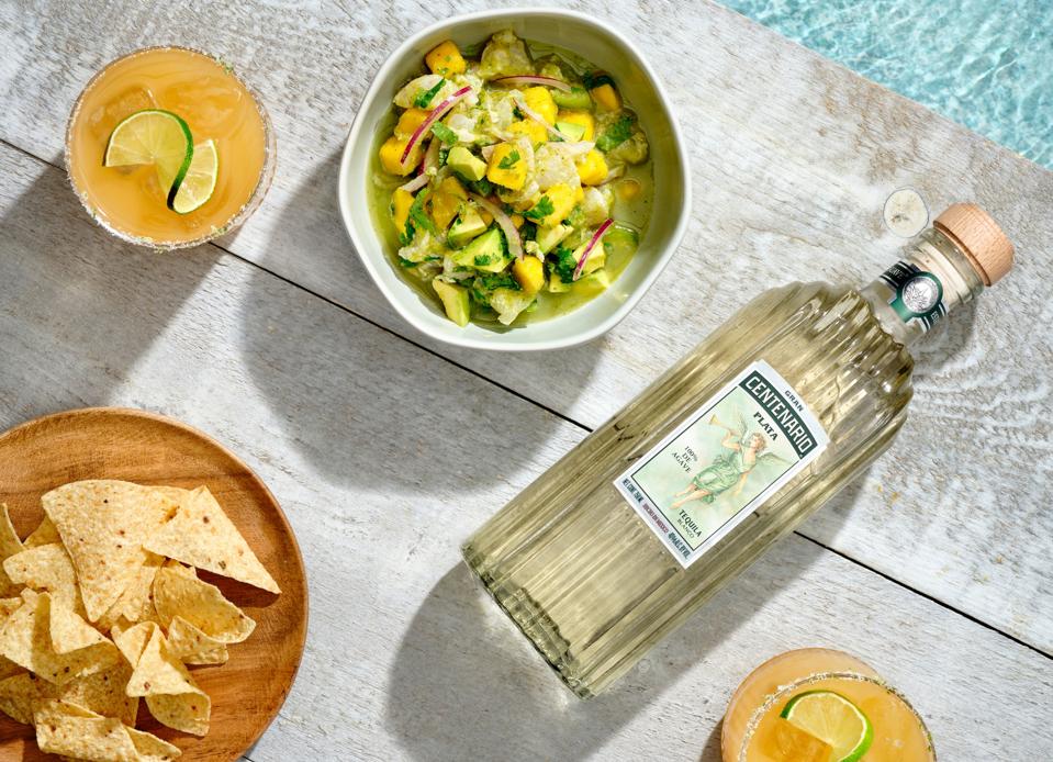 A bowl of ceviche, a bottle of tequila, and a couple cocktails sitting poolside