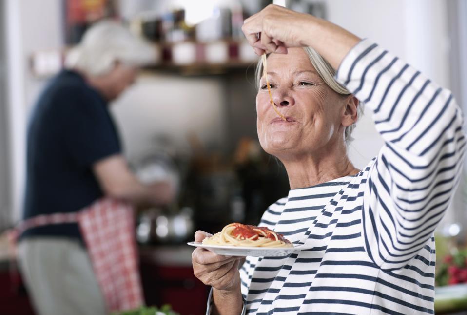 Germany, Wakendorf, Senior woman eating noodles, man cooking in background