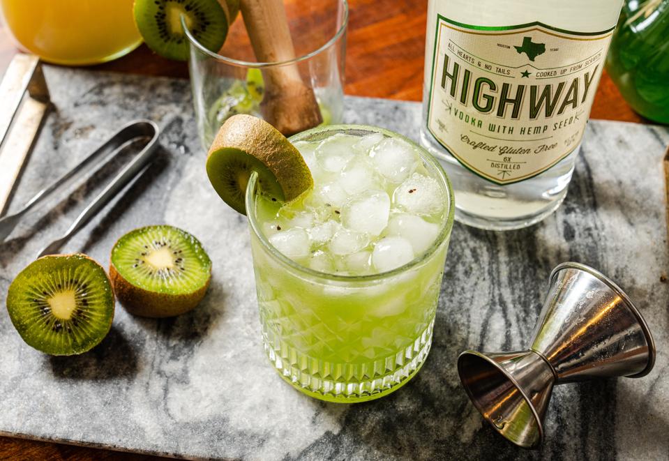 a kiwi sour cocktail with fresh kiwi slices and a bottle of Highway Vodka