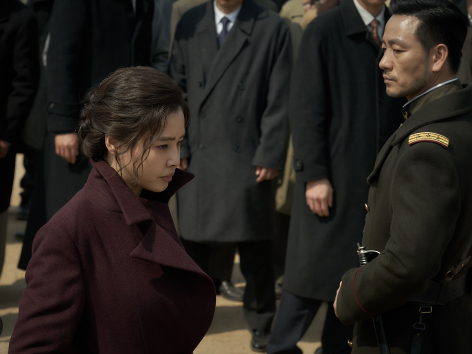 A Korean woman in a dark coat walks past a Japanese officer.