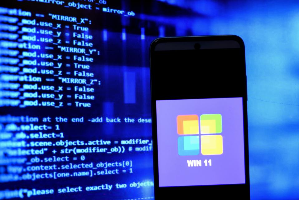 Windows 11 logo on a smartphone, screen behind showing code.