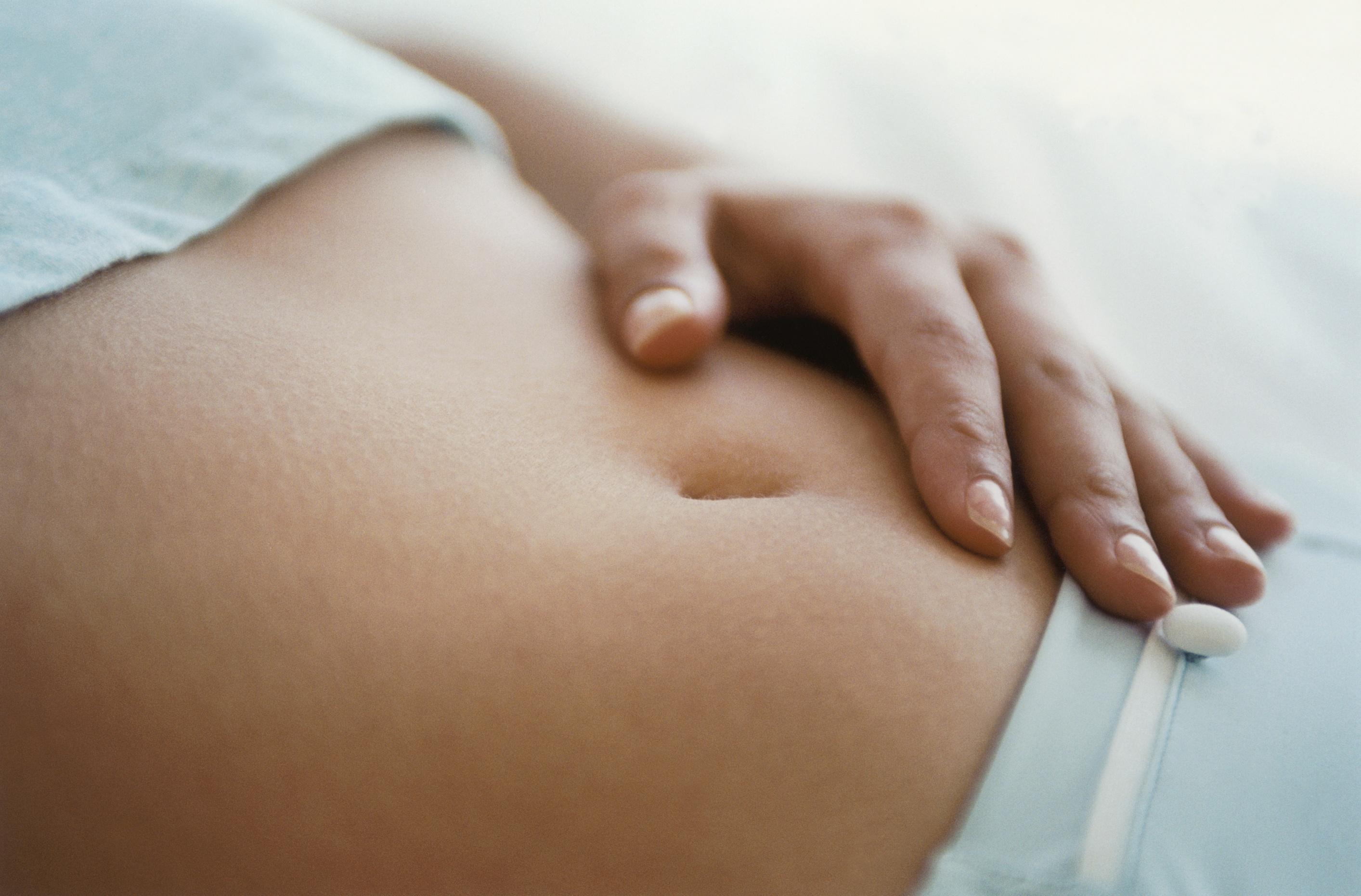 Close-up of a woman's abdomen with a hand resting nearby and a single white pill on bedding.