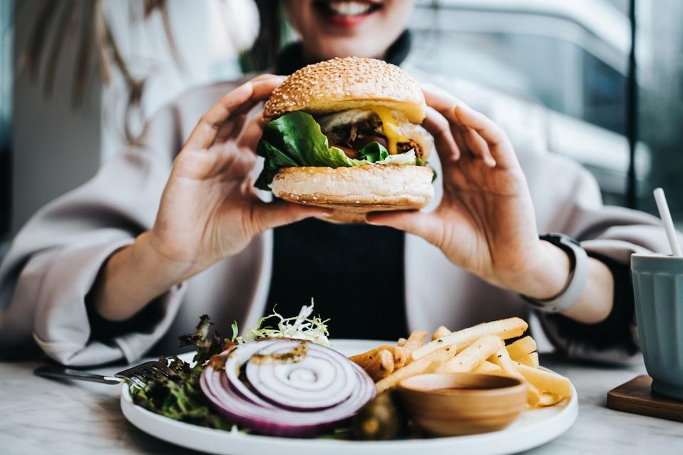 Close up of happy young Asian woman sitting by the window in a restaurant, enjoying lunch during the day. She is having freshly made delicious burger with fries and side salad. Lifestyle, people and food concept