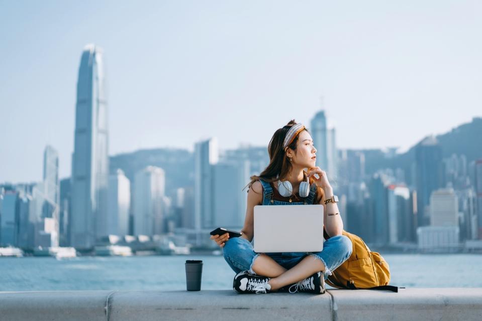 Young Asian woman sitting cross-legged by the promenade, against urban city skyline. She is wearing headphones around neck, using smartphone and working on laptop, with a coffee cup by her side. Looking away in thought. Lifestyle and technology