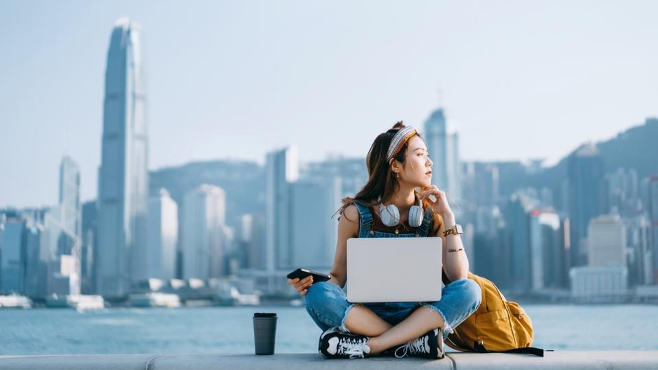 Young Asian woman sitting cross-legged by the promenade, against urban city skyline. She is wearing headphones around neck, using smartphone and working on laptop, with a coffee cup by her side. Looking away in thought. Lifestyle and technology