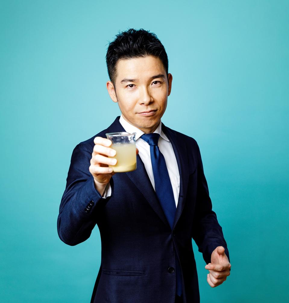 Manabu Aizawa holds a cup of sludge while wearing a suit and tie.