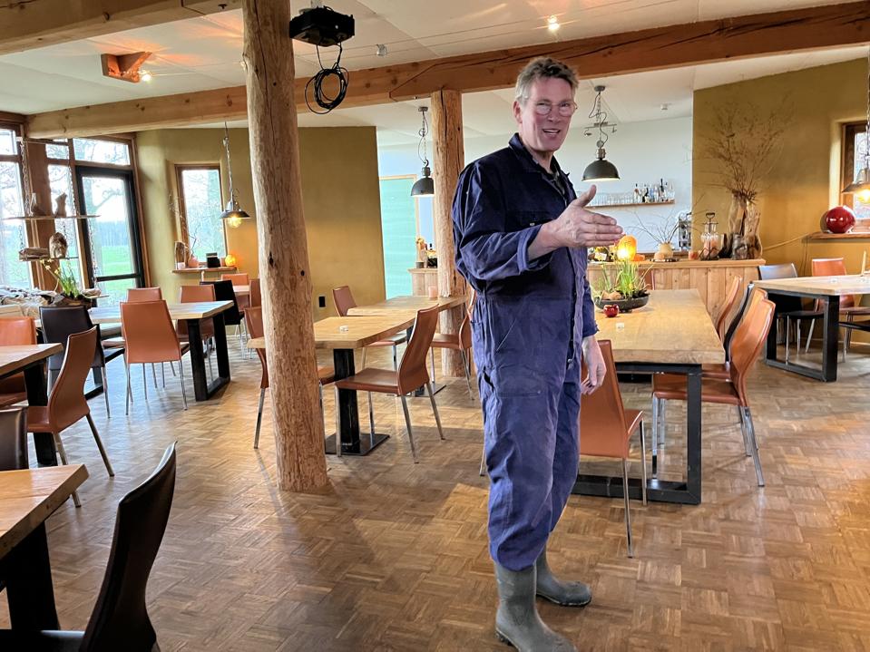 A man in overalls and rain boots stands in a bright room with tables and wooden beams