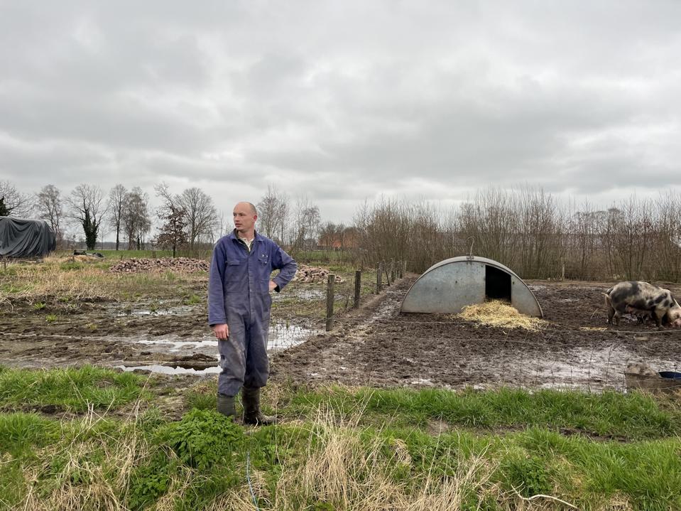 A man in overalls stands outside next to a muddy pen with a big black-and-pink pig