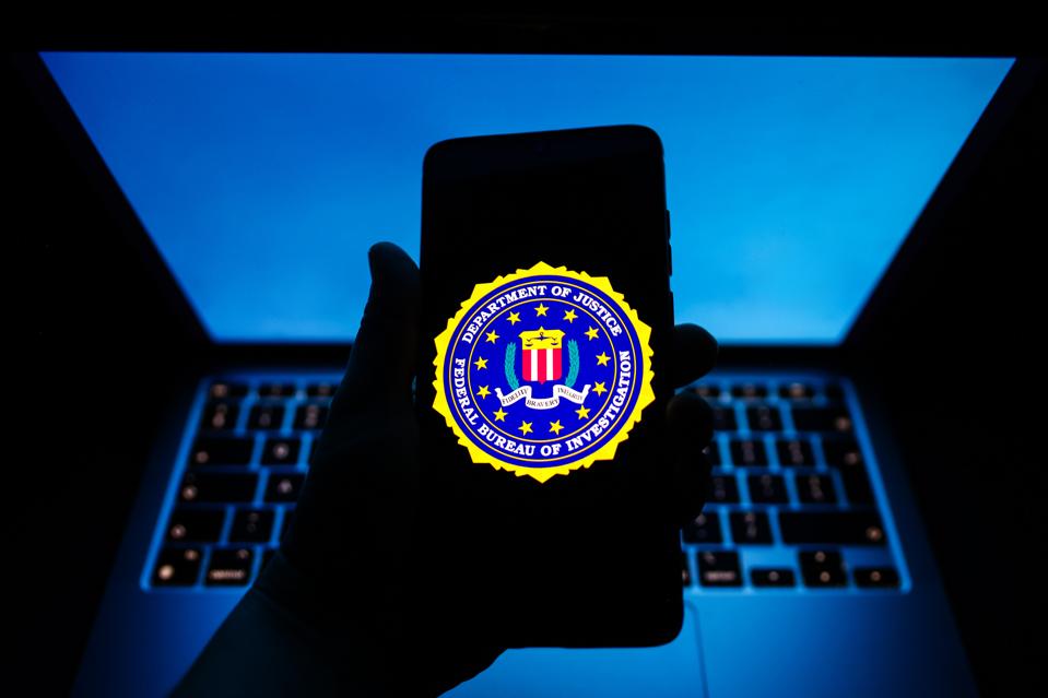 FBI shield seen on a smartphone, in hand, with an open laptop, illuminated in blue, in the background.
