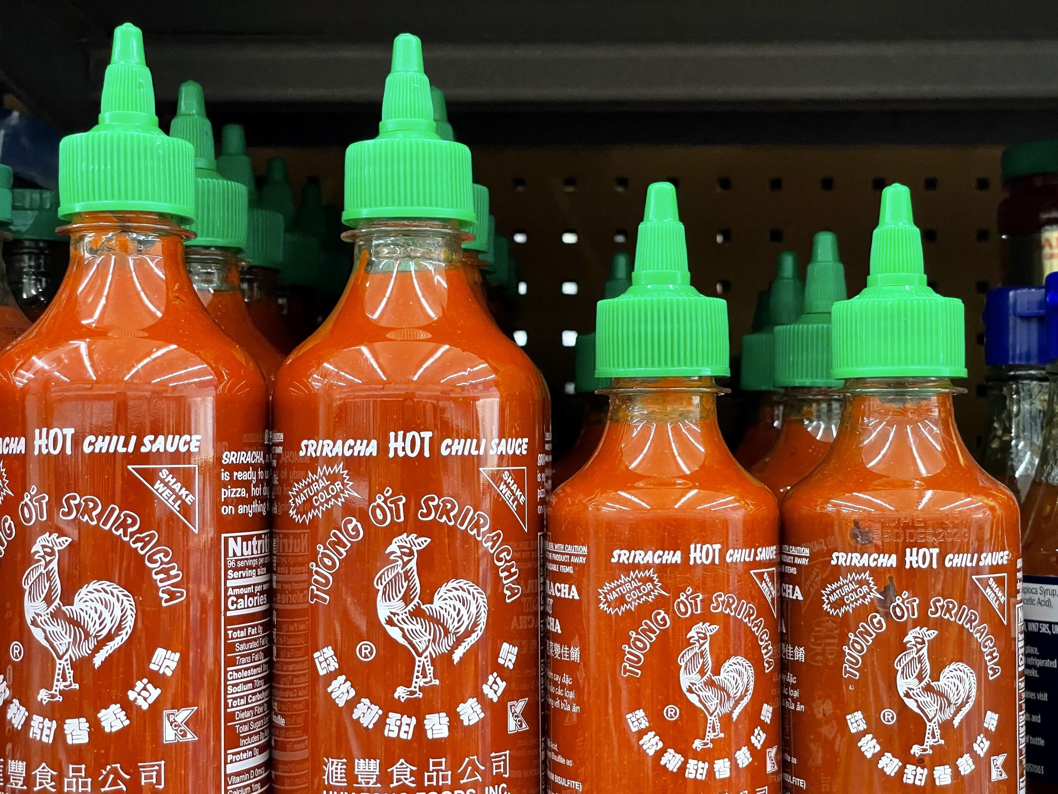 Bottles of Sriracha hot sauce of differing sizes sit on a shelf.