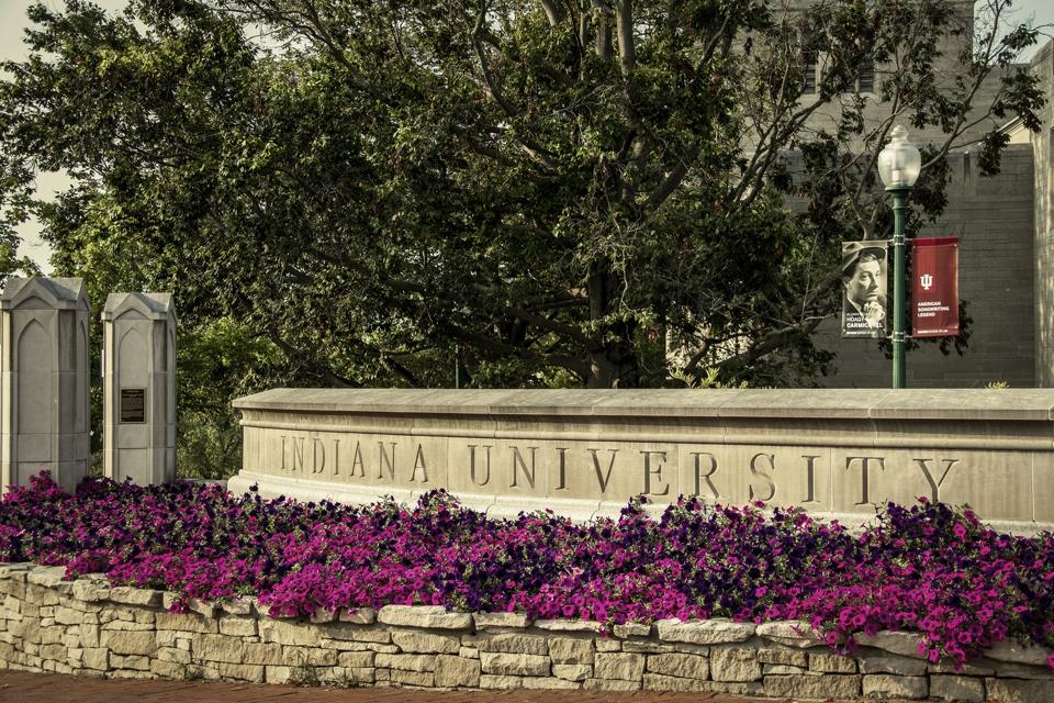 Bloomington USA - Gate into Indiana University with planter full of bright pink flowers and sign on lampost of Alumni Hogy Carmichael American Songwriting Lenand with trees and building in background.