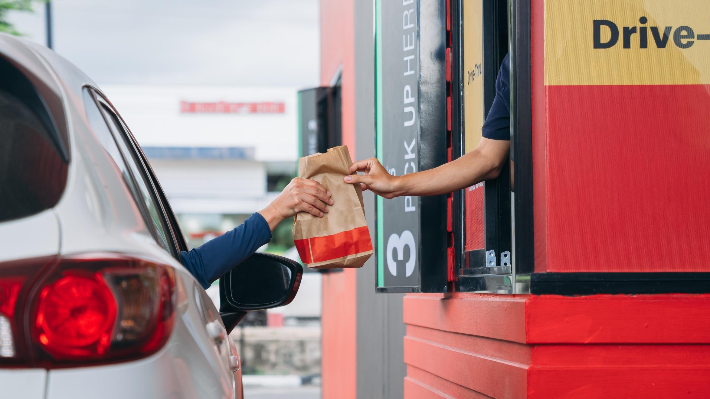 An arm extends from a white vehicle to receive a bag of food from a drive thru.