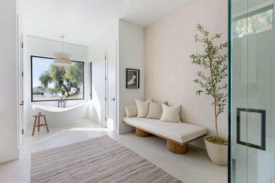 Spacious white-walled bathroom with chaise, freestanding tub, picture window and potted tree.