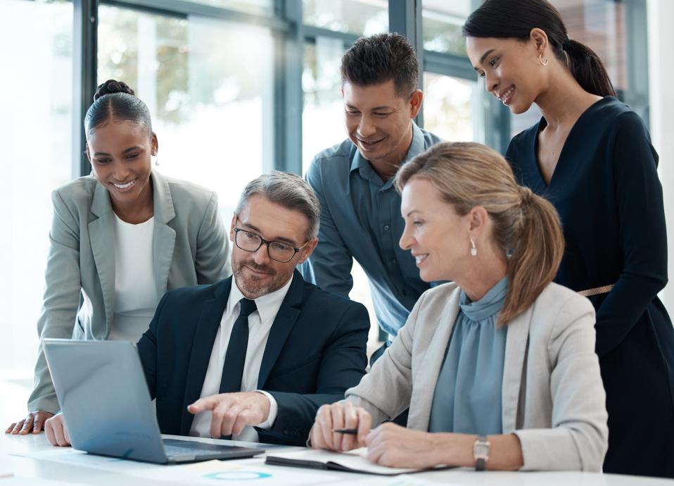 Group of business leaders, male and female, smiling as they’re gathered around a male colleague who is pointing to something on a laptop screen. C-suite enthusiasm for tech tools concept. 