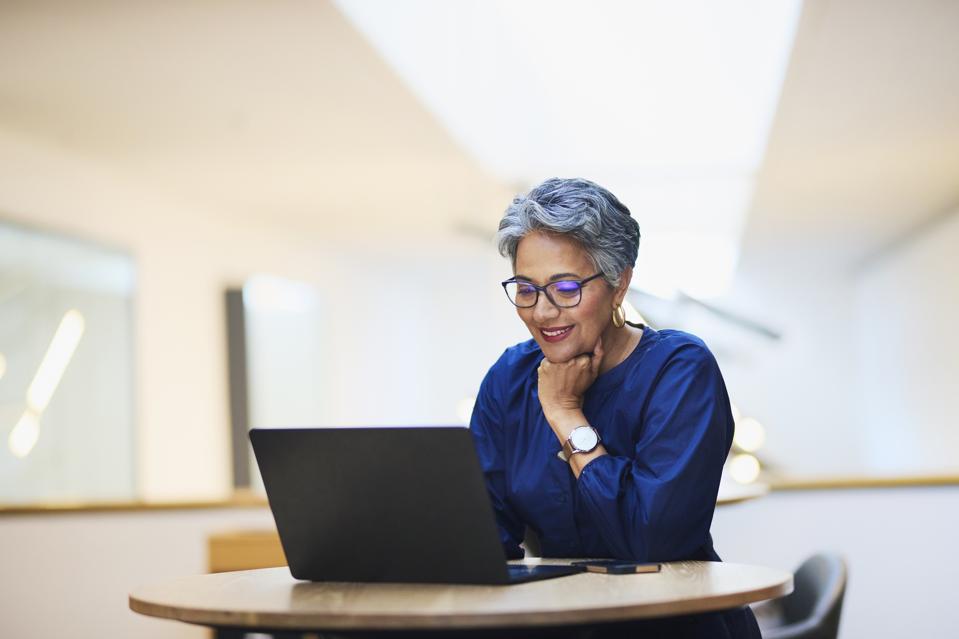 A mature woman with short hair and gray glasses smiles while working on a laptop at a round wooden table. She learns new technological skills.