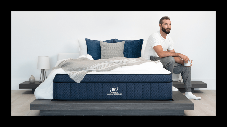 A bearded man sitting on the edge of the Brooklyn Bedding Aurora Luxe Cooling Mattress.