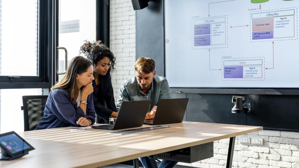 Group of business people working, the team are sitting at a board room table looking at a laptop computer. All dressed in casual business clothing with a window behind them.