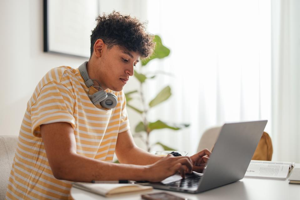 Portrait of a Handsome Teenage Boy Sitting at the Desk and Using a Laptop Computer in His Room at Home