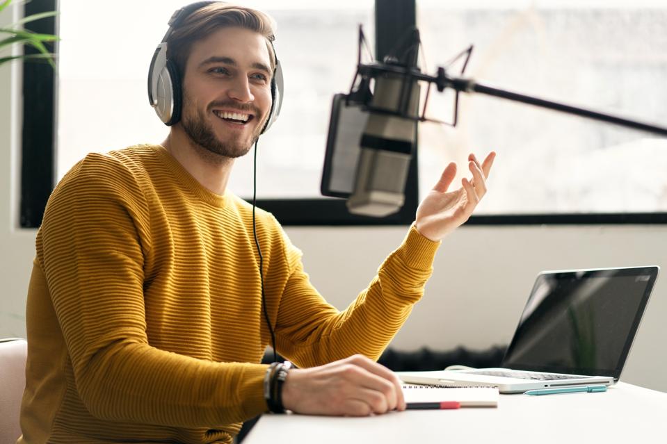Man blogger smiling while broadcasting his live audio podcast in studio using headphones, laptop and headphones