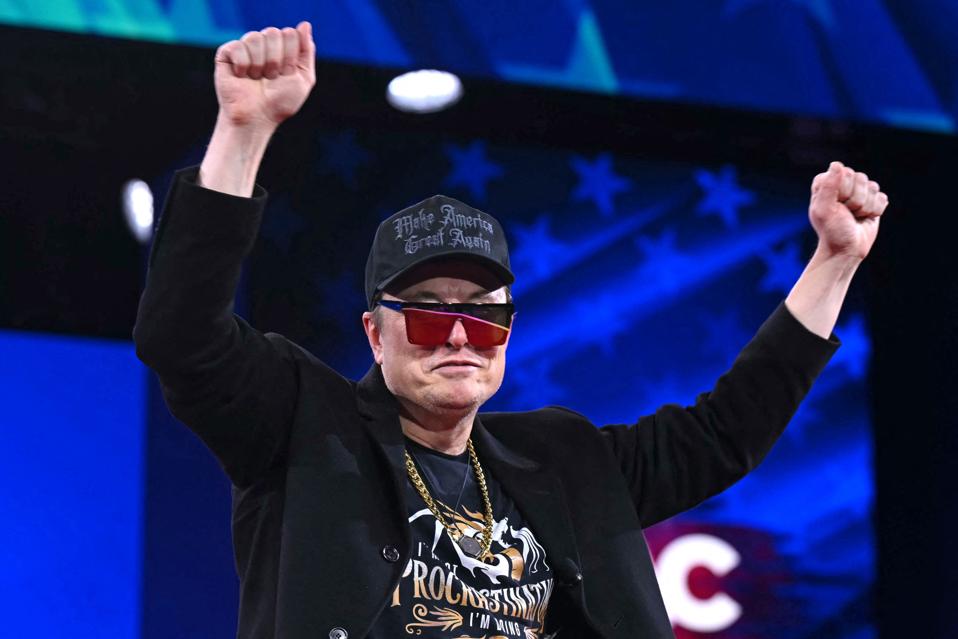 Elon Musk on stage wearing sunglasses and a trucker hat. 