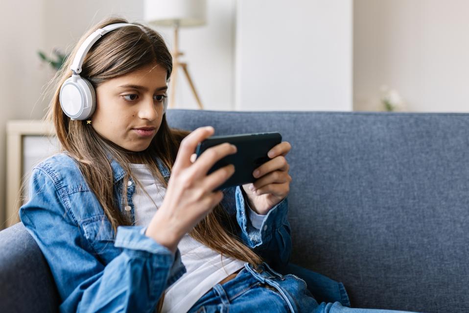 Child teen girl with headphones playing on mobile game using phone app. Concentrated teenager female gamer player using smartphone relaxing on sofa at home. Addicted to social media and technology concept.