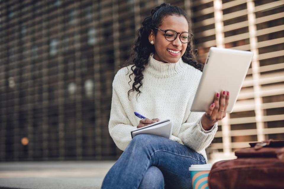 A young black woman in a white sweater sits on a bench in front of an apartment complex and works on a tablet