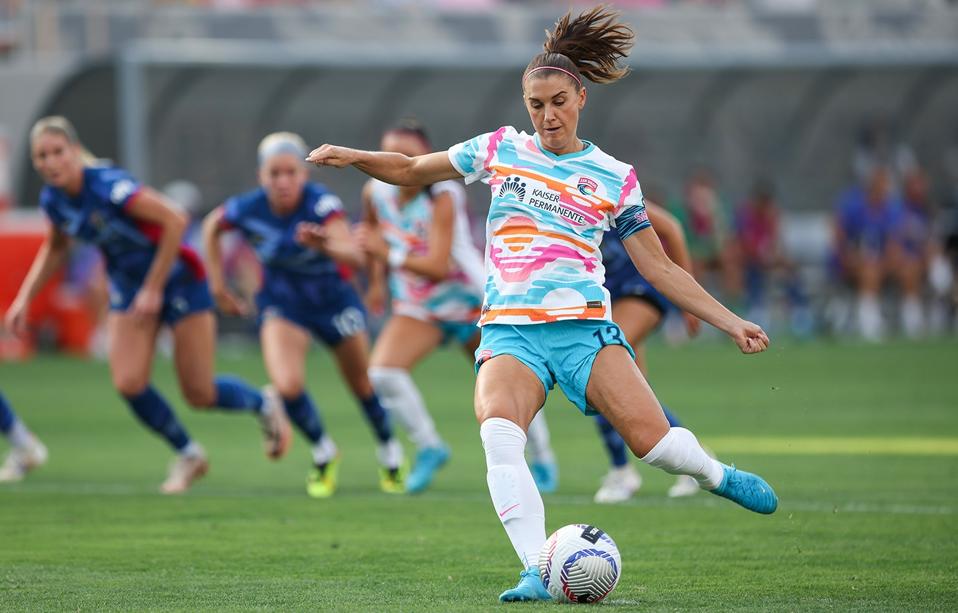Alex Morgan wearing a San Diego Wave kit while leaning back and kicking the ball during a soccer game.
