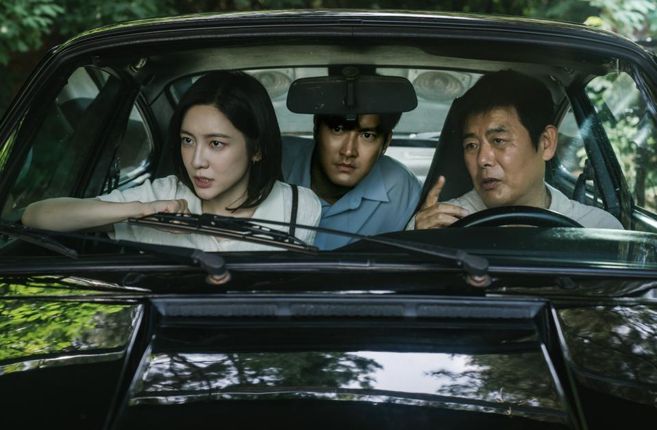 Two men and a woman sit in a car discussing someone outside.