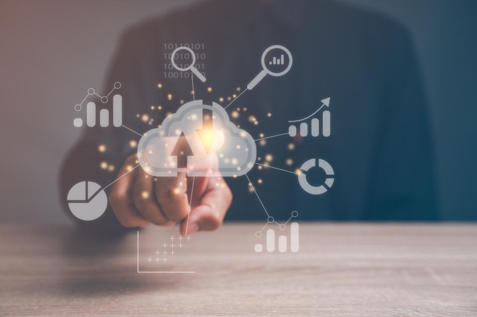 Digital image of cloud surrounded by business charts and graphs. From behind, a person reaches forward to touch the cloud image with a forefinger.