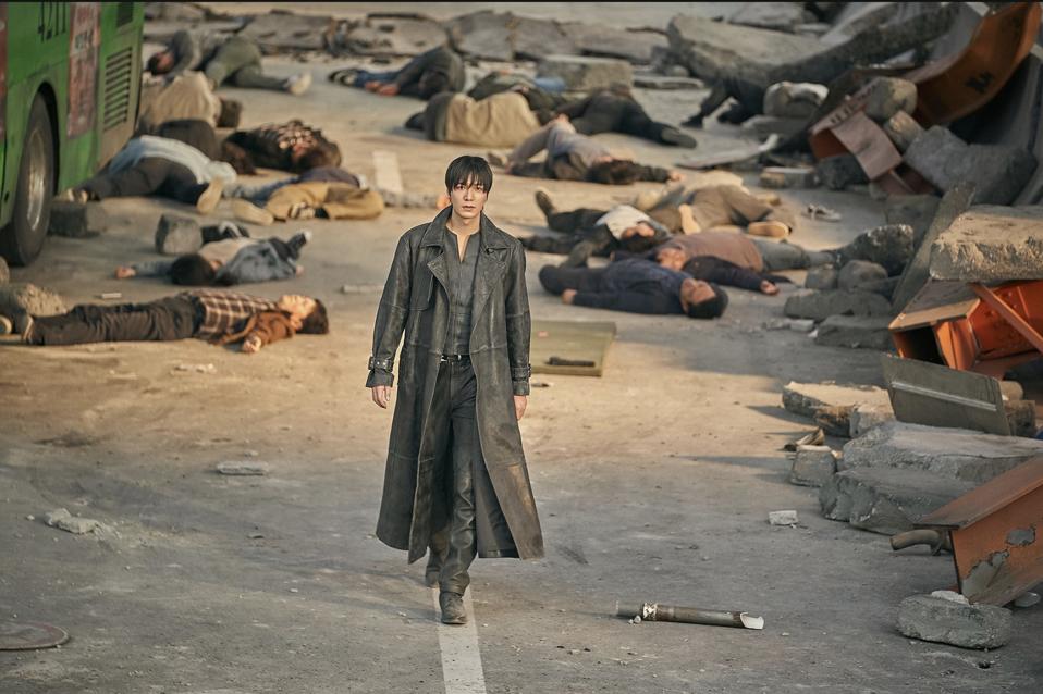 A man with a long leather coat walks down a street littered with bodies.
