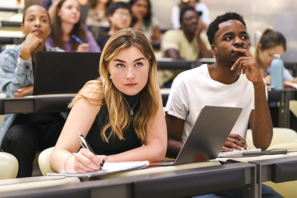 Young female student writing in book while sitting by male friend at university classroom